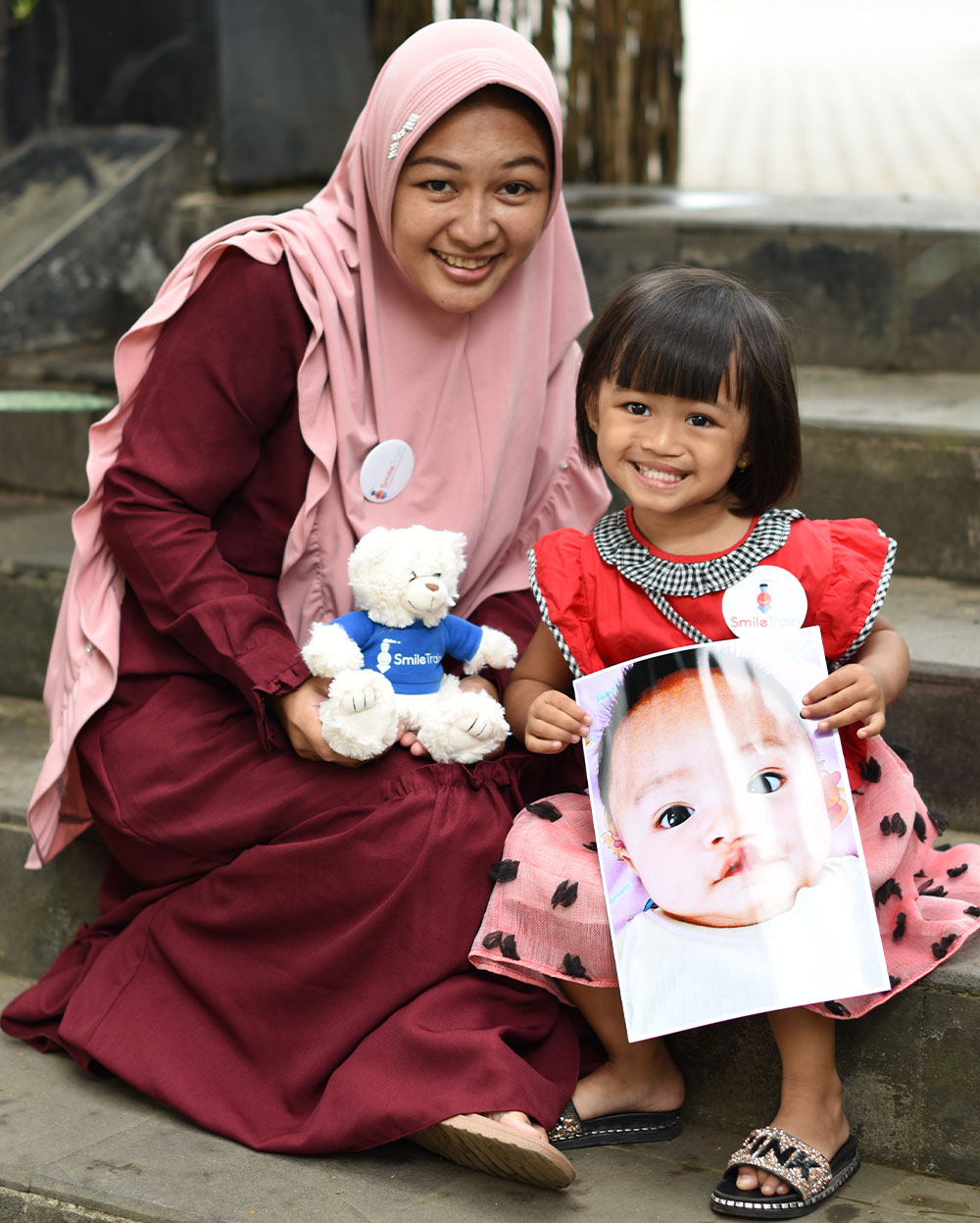 Khalisa smiling with her mother whilst holding a picture of herself before cleft surgery