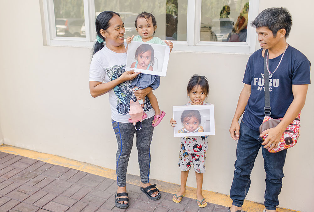 Ernie and Ermalen after Smile Train-sponsored cleft surgery holding pictures of themselves before surgery, flanked by their parents