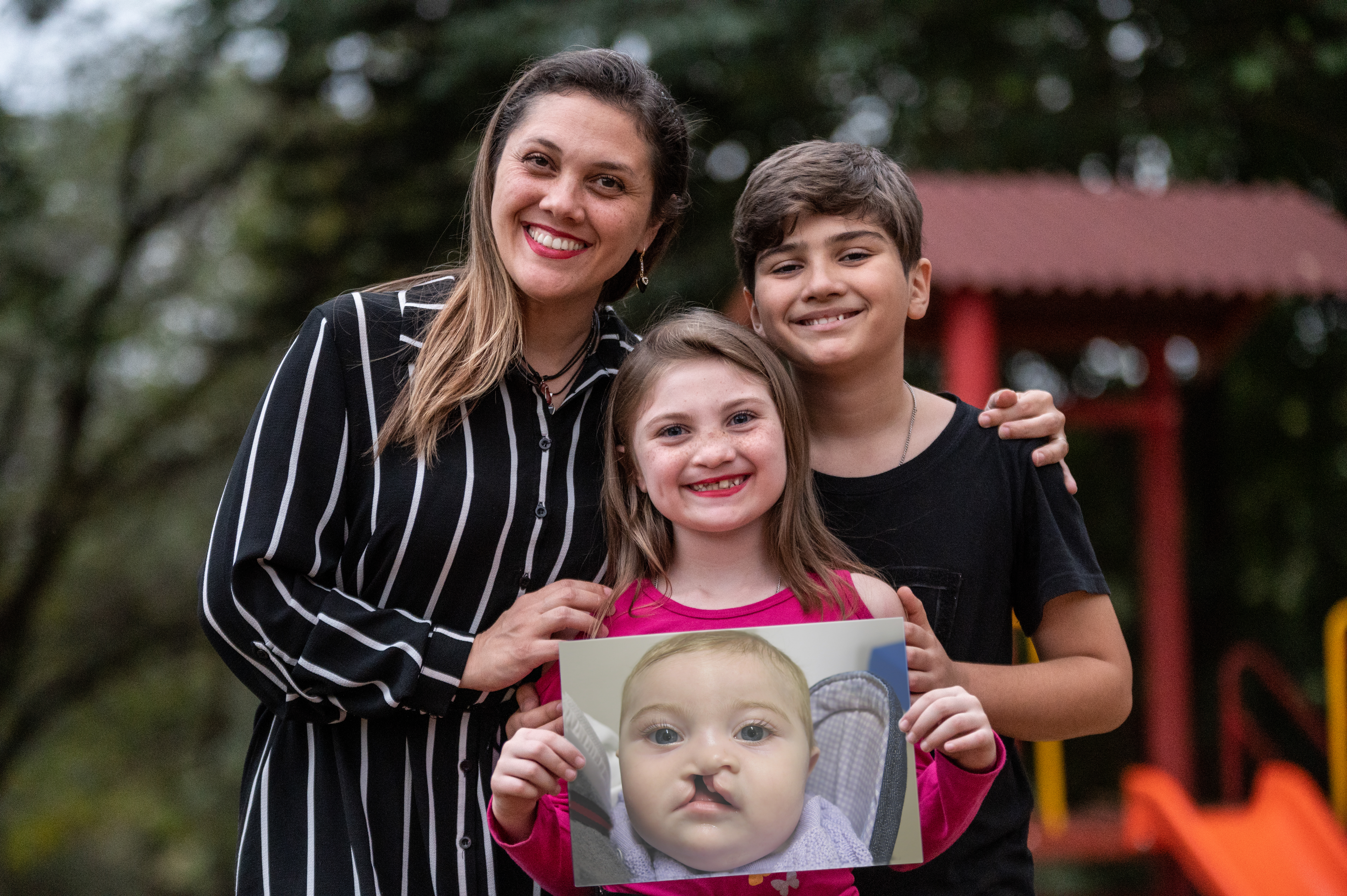Grazi and Nilo standing behind Milena with their hands on her shoulders. Milena is holding a photo of herself as a baby before cleft surgery