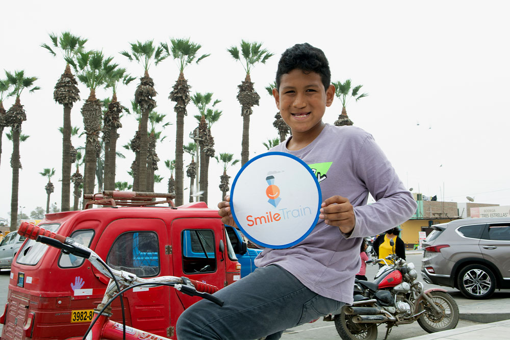 Imanol sitting on his bike in front of a street. He’s holding a Smile Train sign