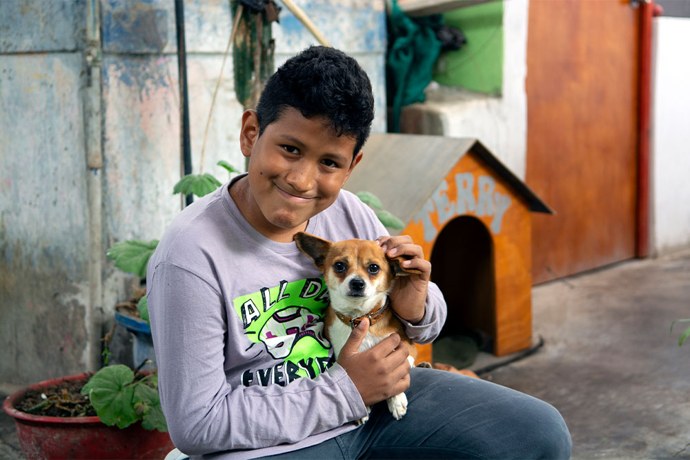 Imanol sitting and holding his pet chihuahua in front of his doghouse